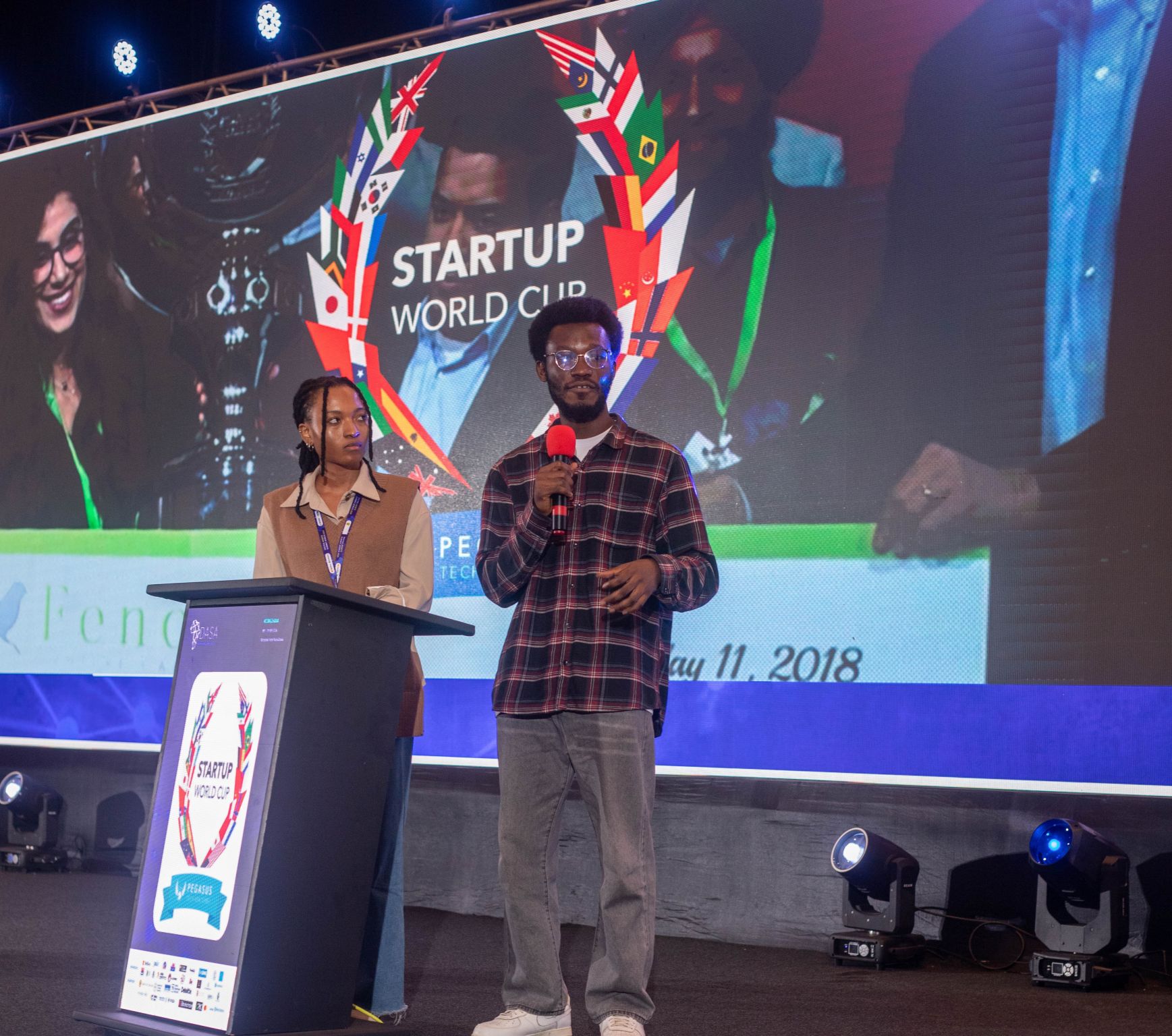 Two presenters stand beside a podium in front of Startup World Cup branding.
